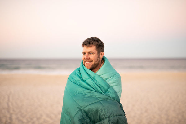 Mint green down puffy blanket wrapped around man on beach, shown in use outdoors
