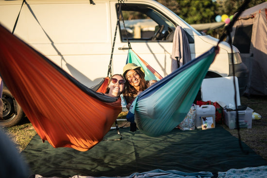 Nakie hammock set up between vehicles at a festival-style campsite during sunset.
