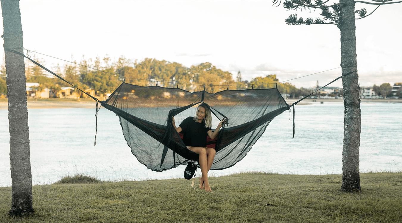 Woman in a Nakie hammock with a bugnet relaxing by the water.