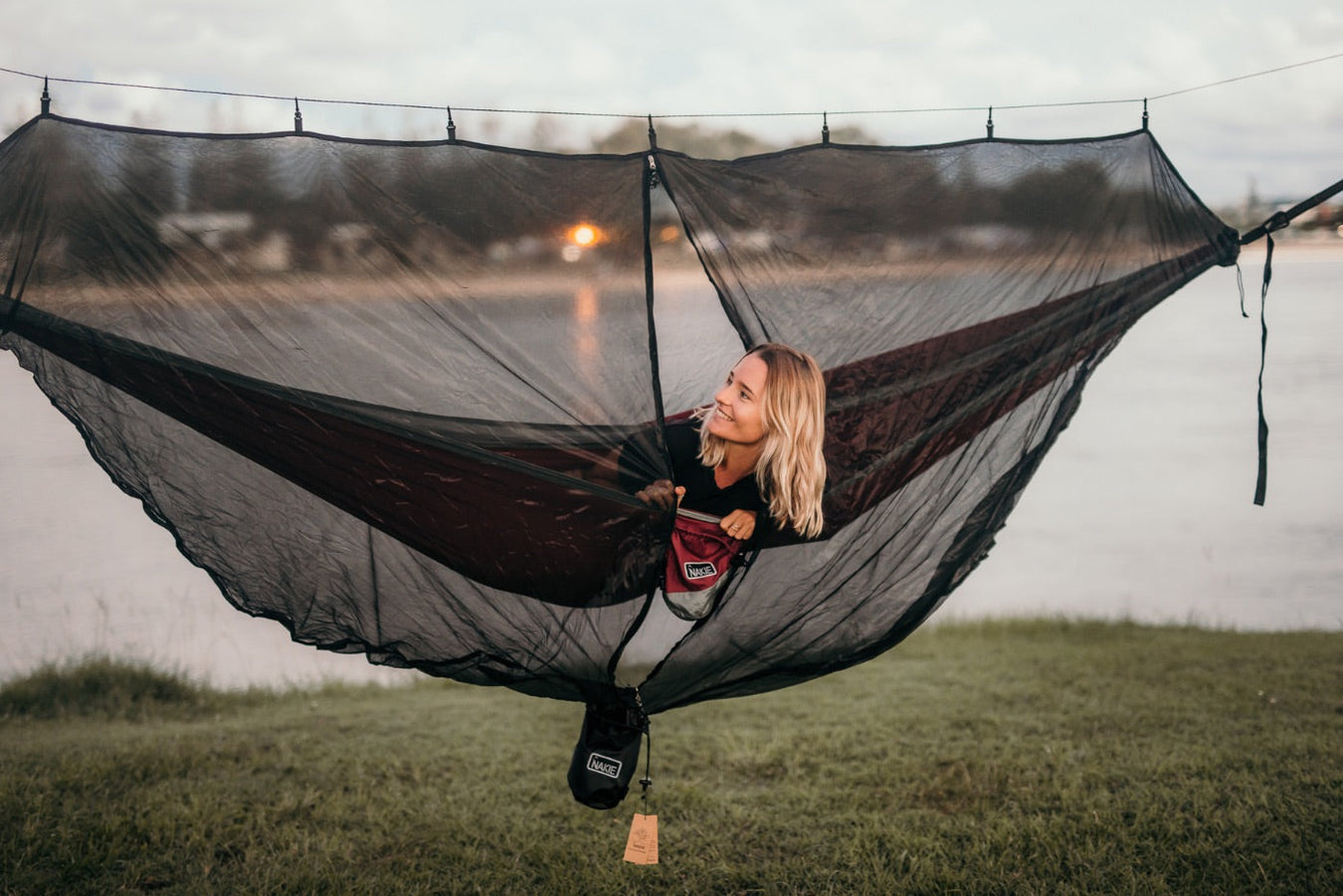 Woman in a Nakie hammock with a bugnet relaxing by the water.