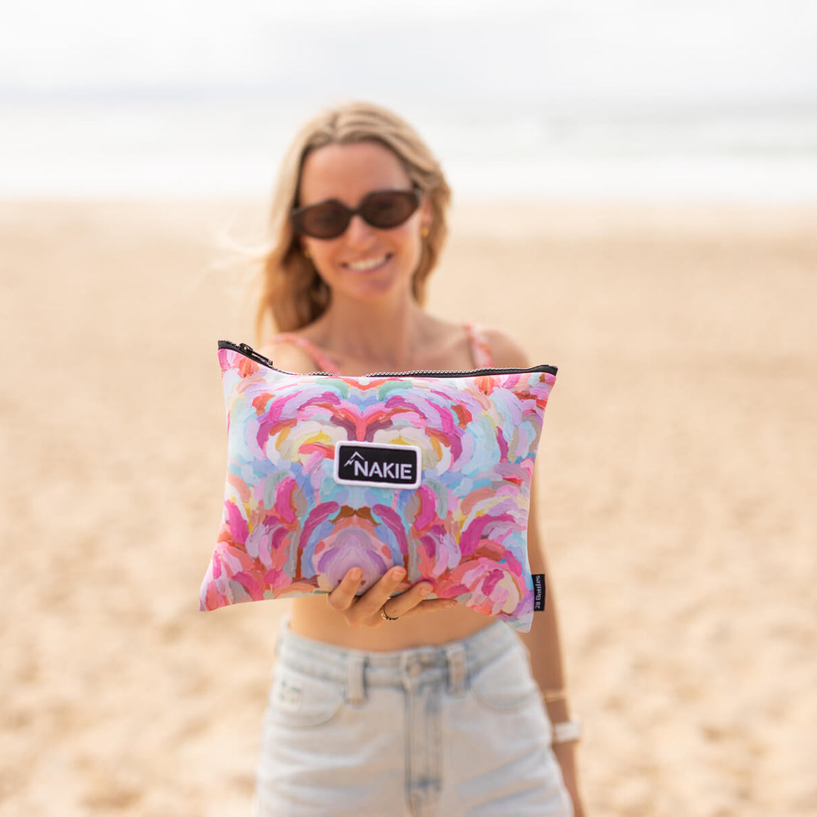 Recycled sand-free beach towel in pink and purple abstract floral print with dark trim, held by woman on beach.