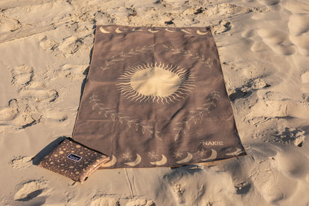 Brown recycled beach towel with celestial sun and moon design, laid flat on sand with matching pouch