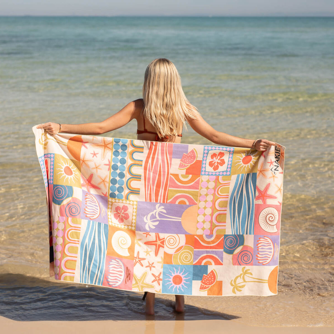 Recycled sand-free beach towel with colorful retro patchwork pattern held open by woman at the shore.