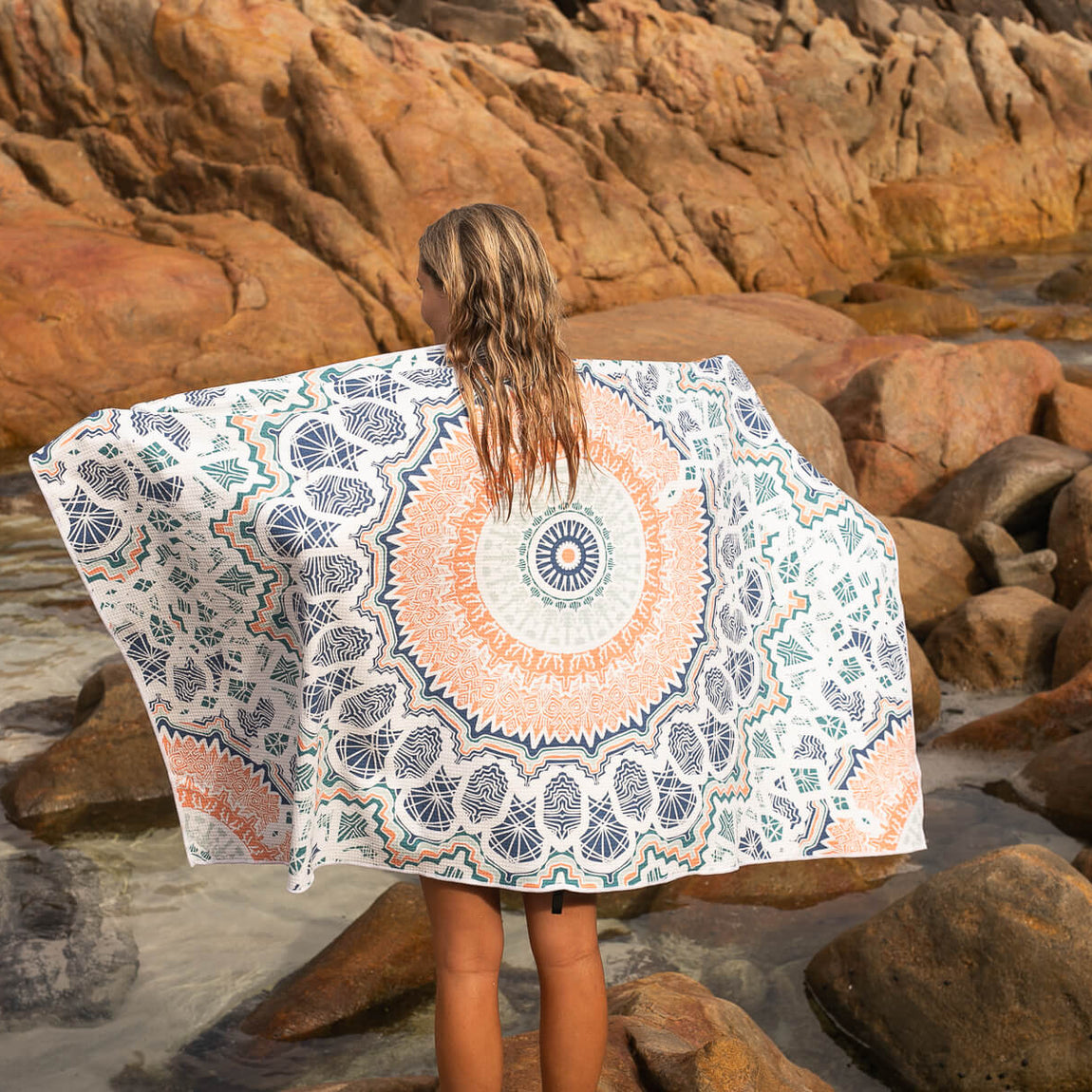 Recycled sand-free beach towel with blue and orange geometric mandala pattern, worn by person at rocky beach