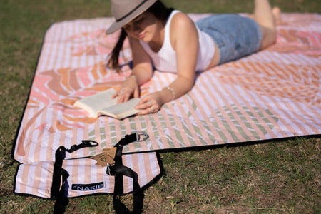 A woman reading a book while lying down on Nakie's recycled picnic blanket, enjoying relaxation and comfort outdoors.